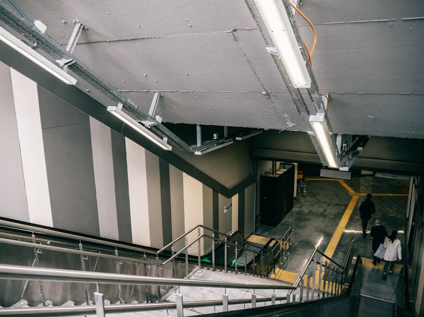 View of a staircase and escalator inside a modern indoor space near Stockwell Station, with several people walking on the tiled floor. The staircase has metal handrails and yellow tactile paving at th