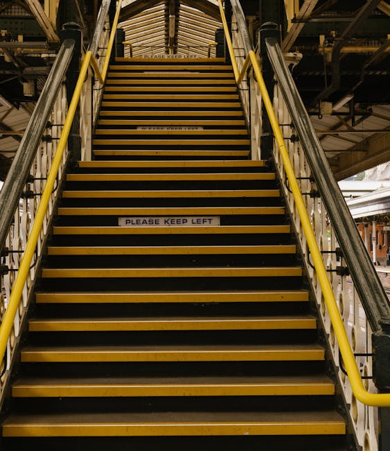 View of a staircase and escalator inside a modern indoor space near Stockwell Station, with several people walking on the tiled floor. The staircase has metal handrails and yellow tactile paving at th